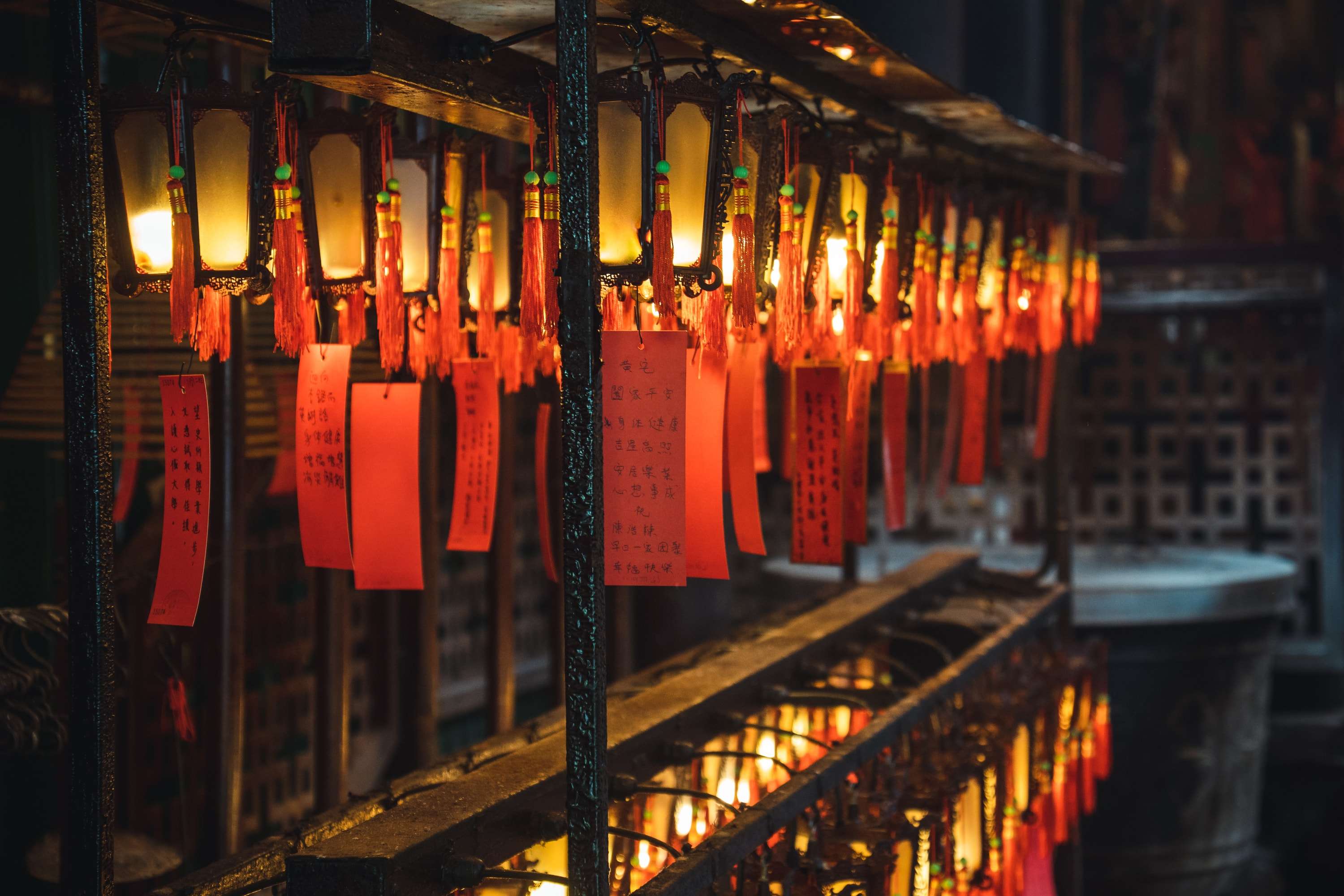 Rows of small lit lanterns with red tassels and red paper prayer tags hang from a dark metal frame inside a temple.