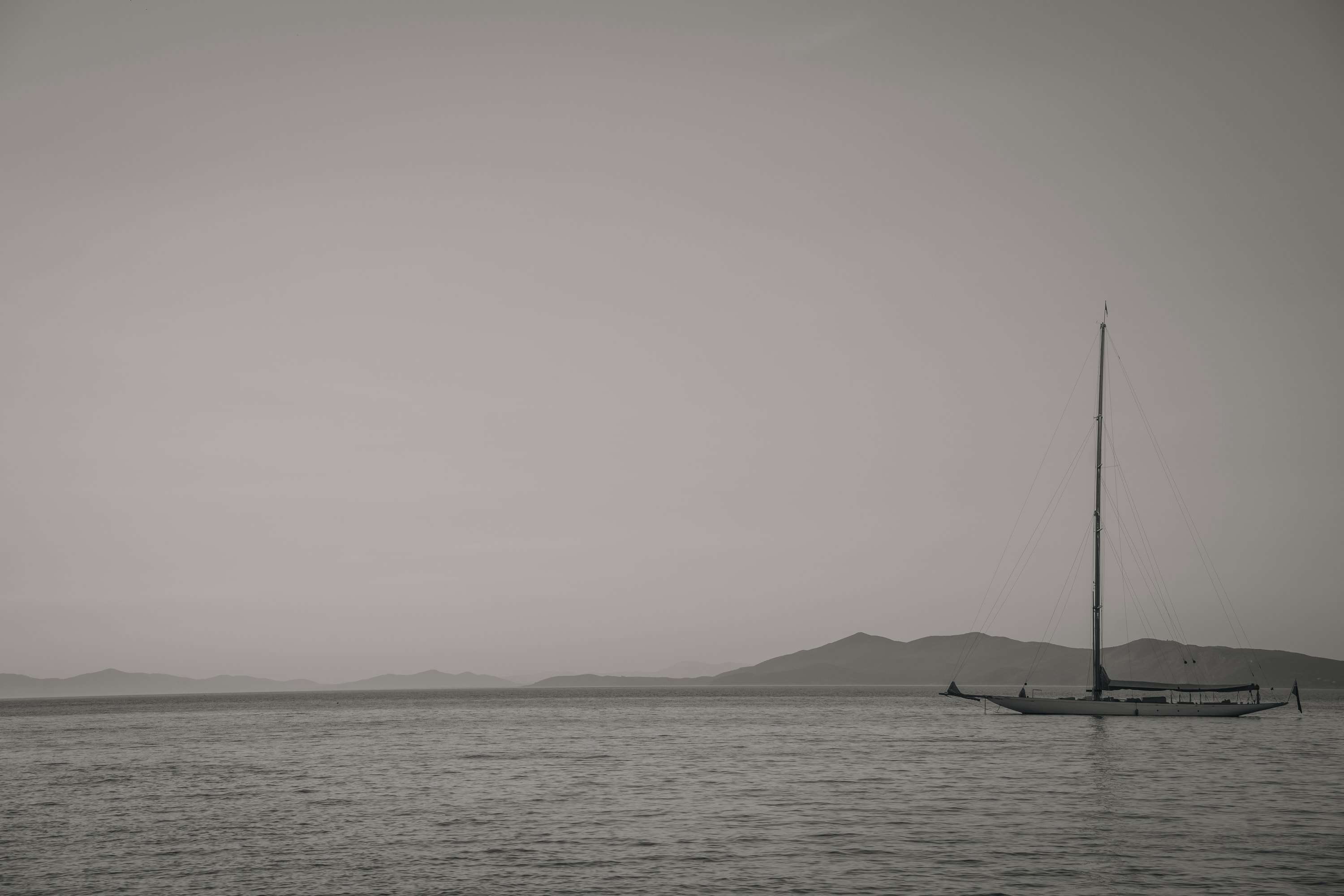 A classic sailing yacht with a tall bare mast lies at anchor on a calm grey sea, with a line of hazy hills and islands behind it.