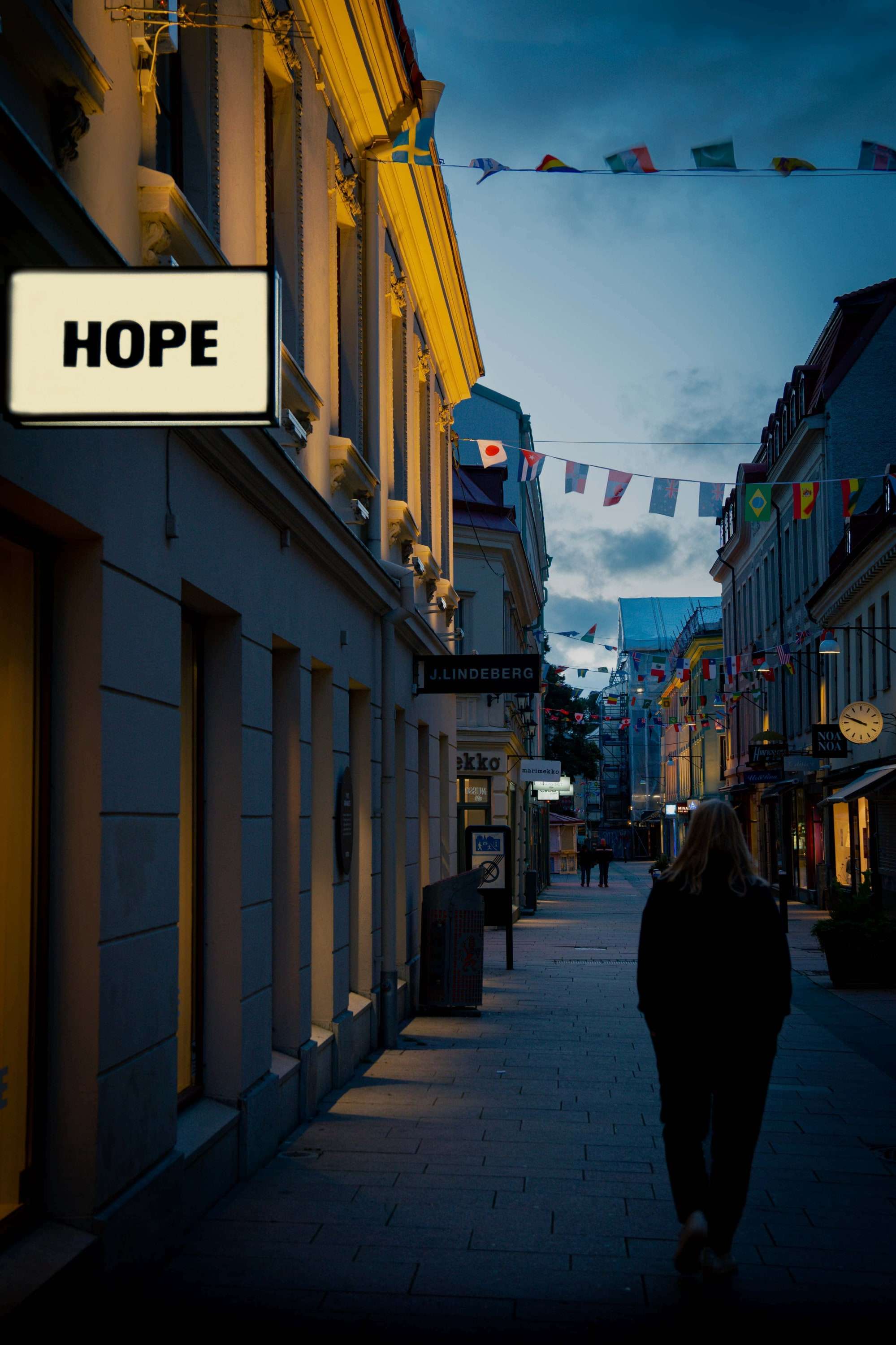 A person walks down an evening street past a lit sign reading HOPE, with bunting strung between the buildings.