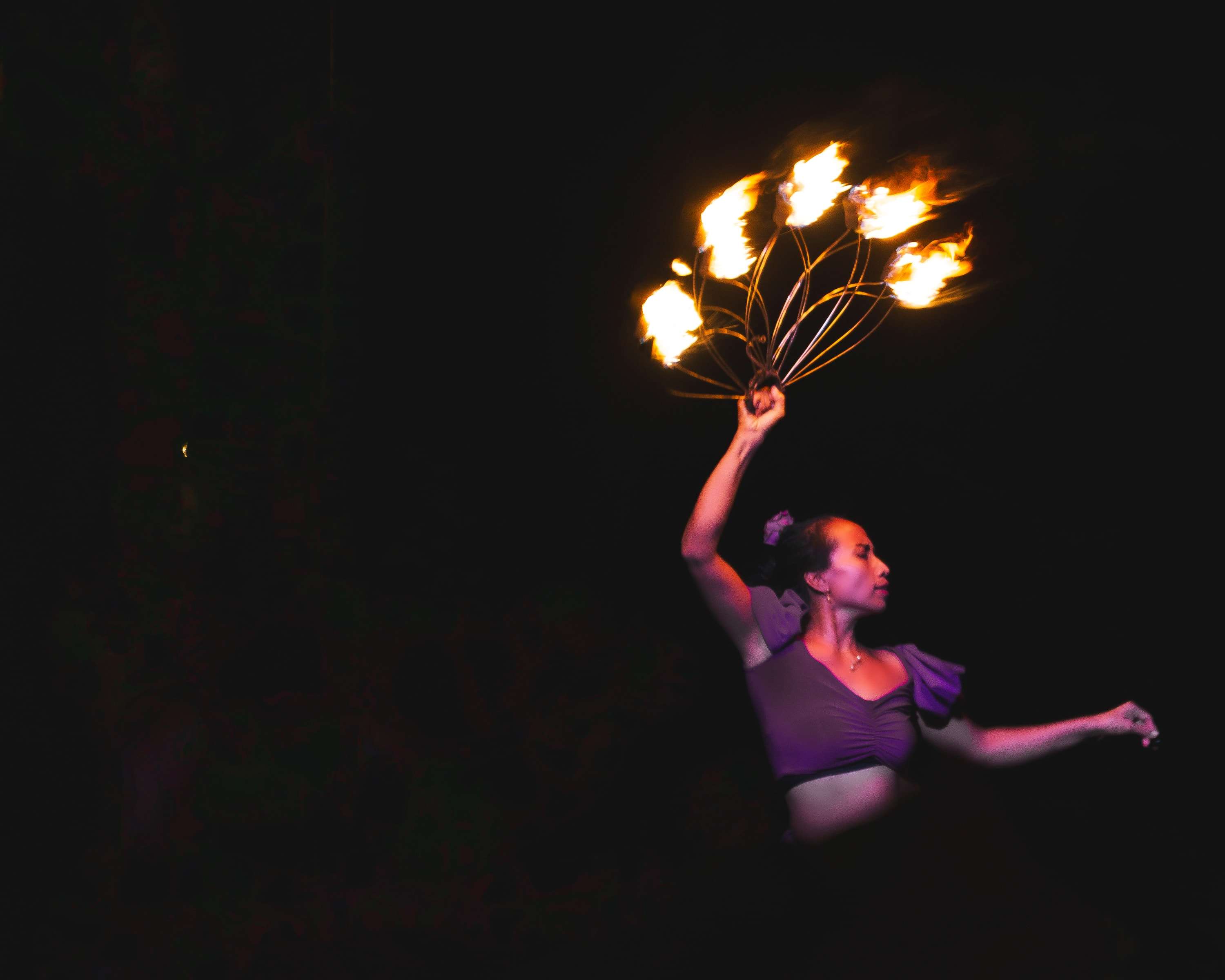 A fire dancer in a purple top turns in profile, one arm raised holding a burning fan of wicks, the other stretched out behind her.