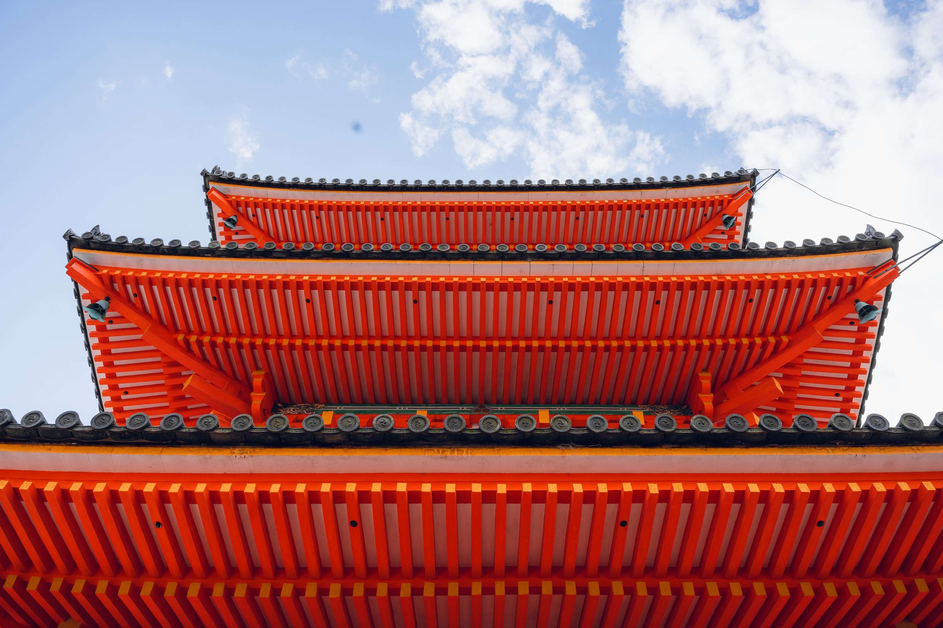 The vermilion roof tiers of a pagoda seen from directly below, stacked against a blue sky with white clouds.