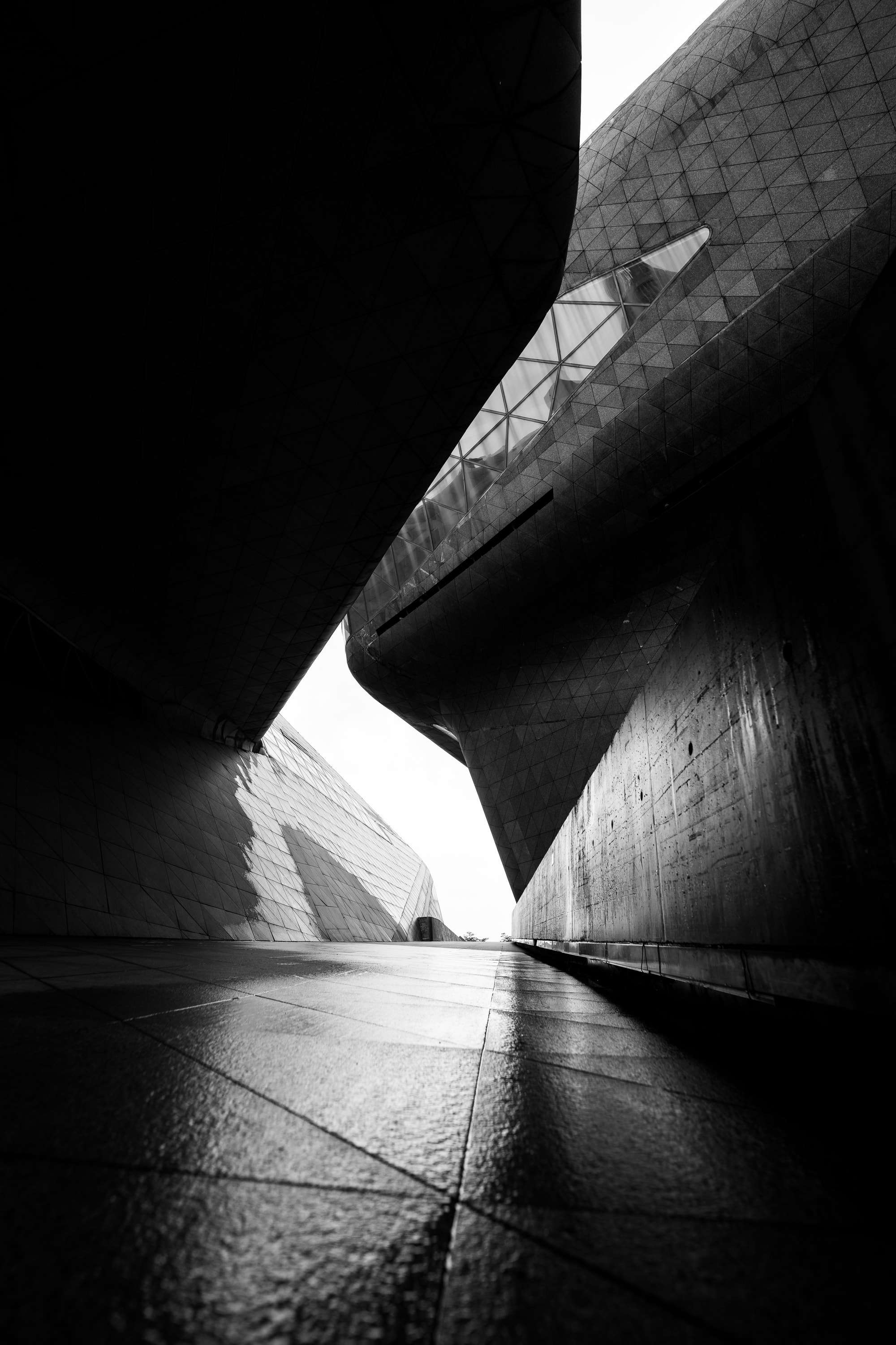 Black and white: a paved walkway runs between the curved, faceted walls of the Guangzhou Opera House toward a bright gap of sky.