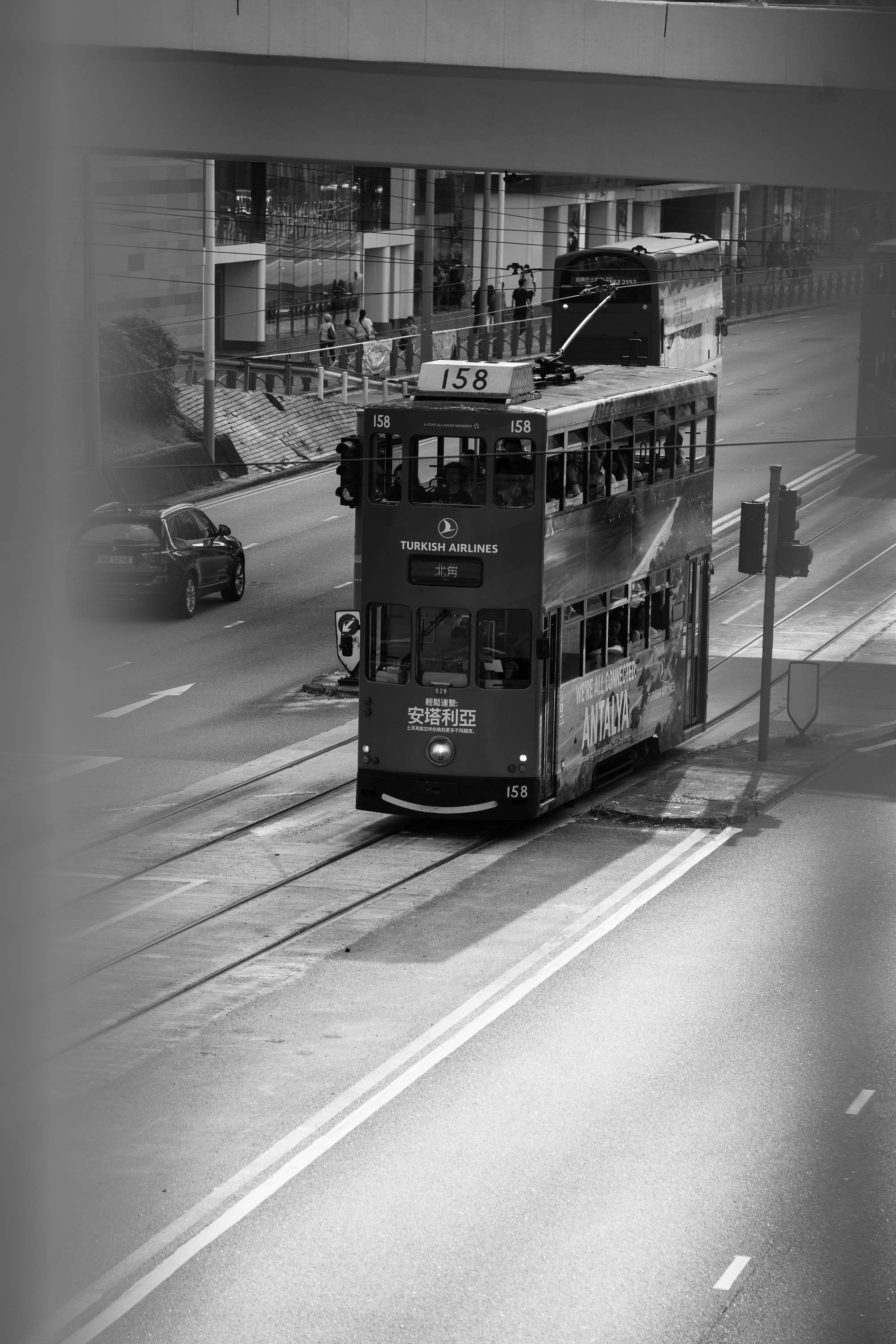 Black and white: double-decker tram number 158, bound for North Point, runs along the tracks under a concrete walkway, a car and a bus behind it.