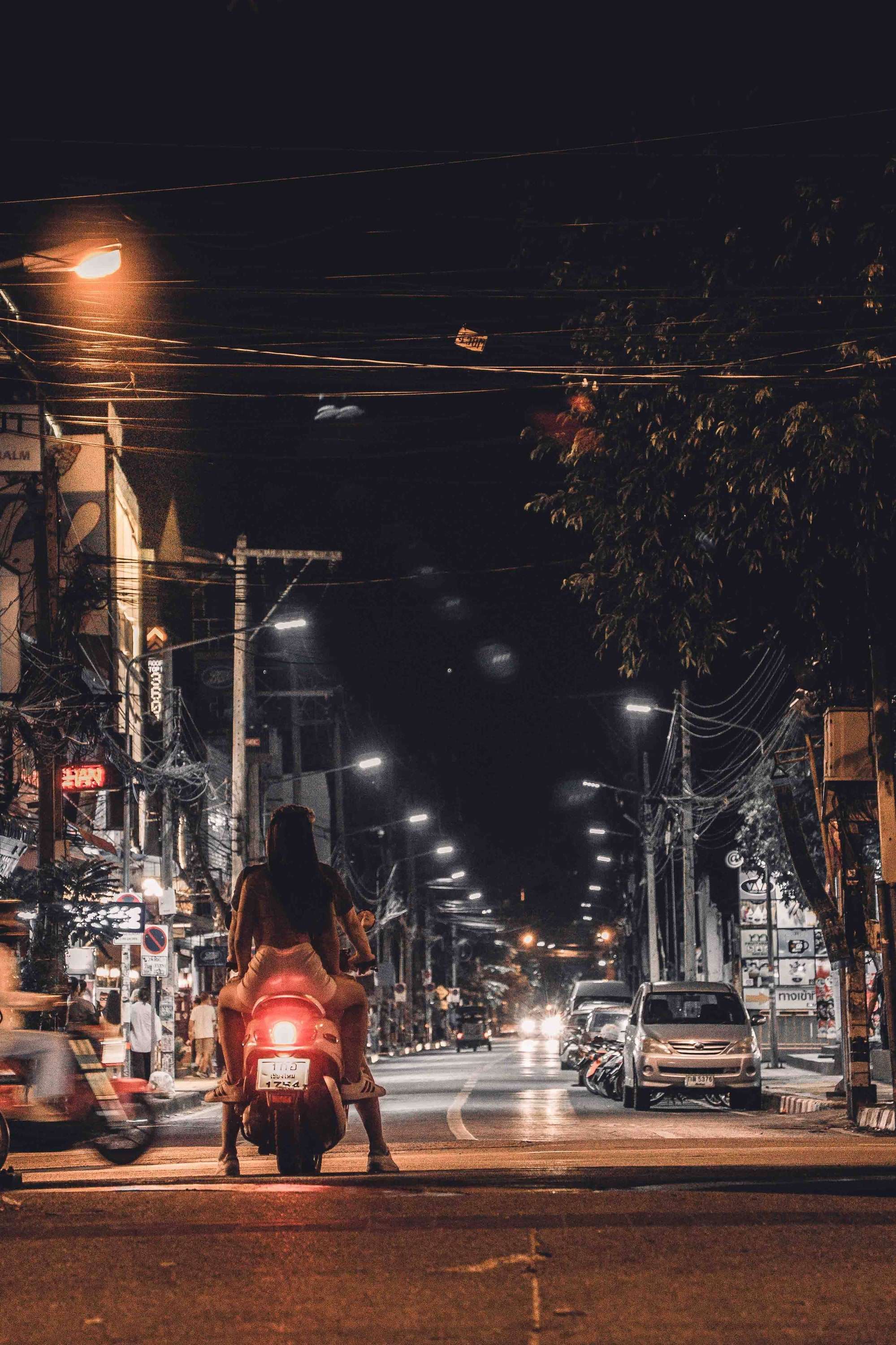 A scooter rider waits at a junction under street lights and tangled overhead cables on a Chiang Mai street at night.