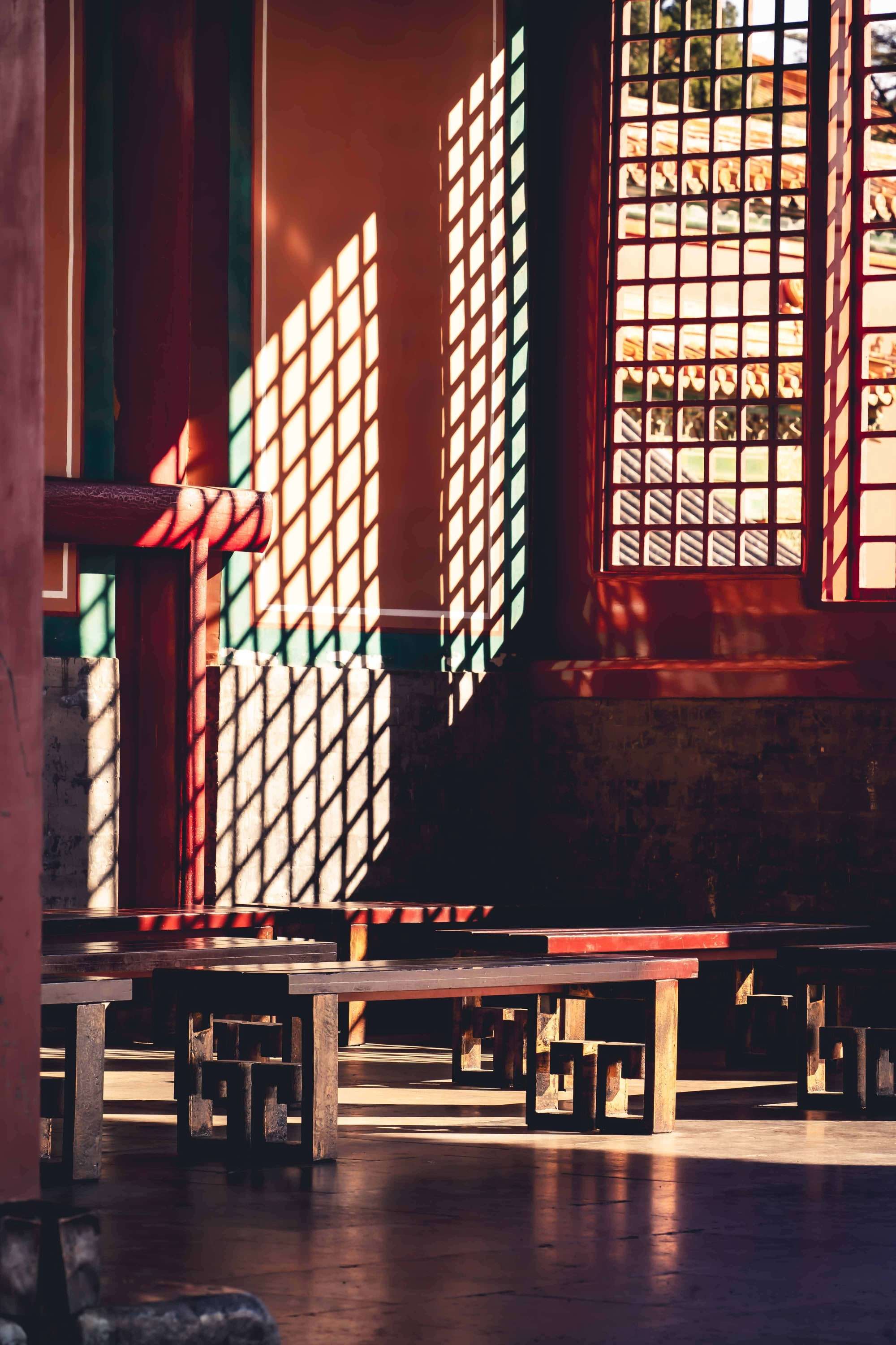 Afternoon light falls through a carved lattice window across dark wooden benches inside a traditional hall.