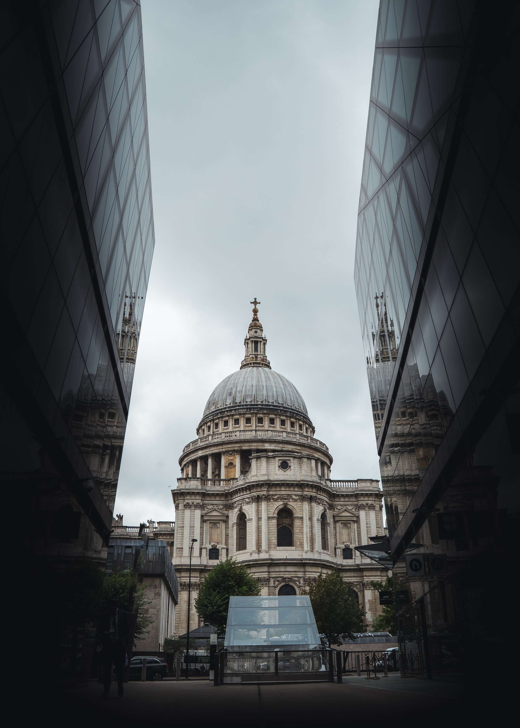 The dome of St Paul's Cathedral seen through a narrow gap between two dark glass walls of a modern building, under a white overcast sky.