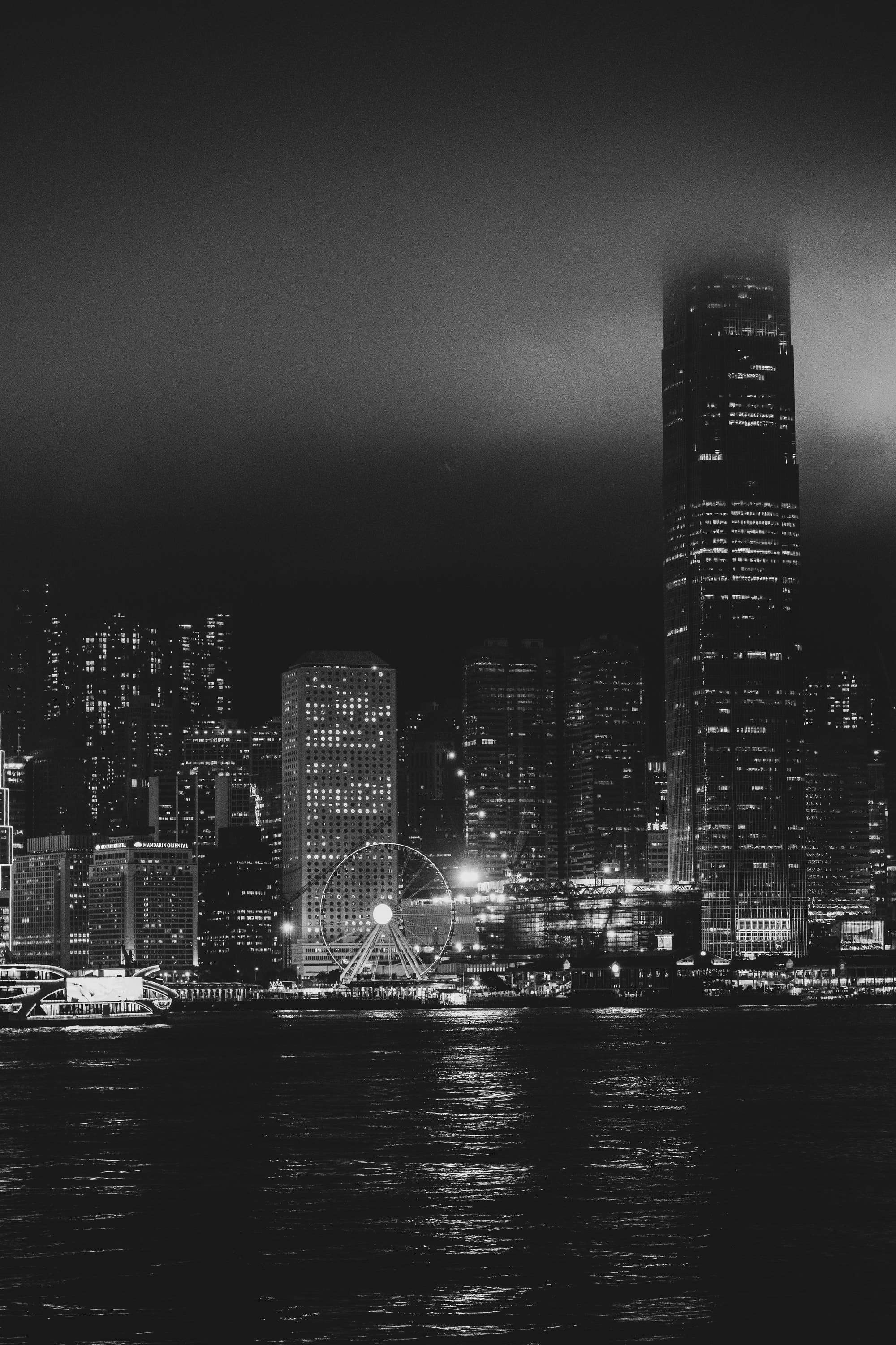 Black and white: the IFC tower disappearing into low cloud above the lit skyline of Hong Kong Island and the Ferris wheel, reflected in dark water.