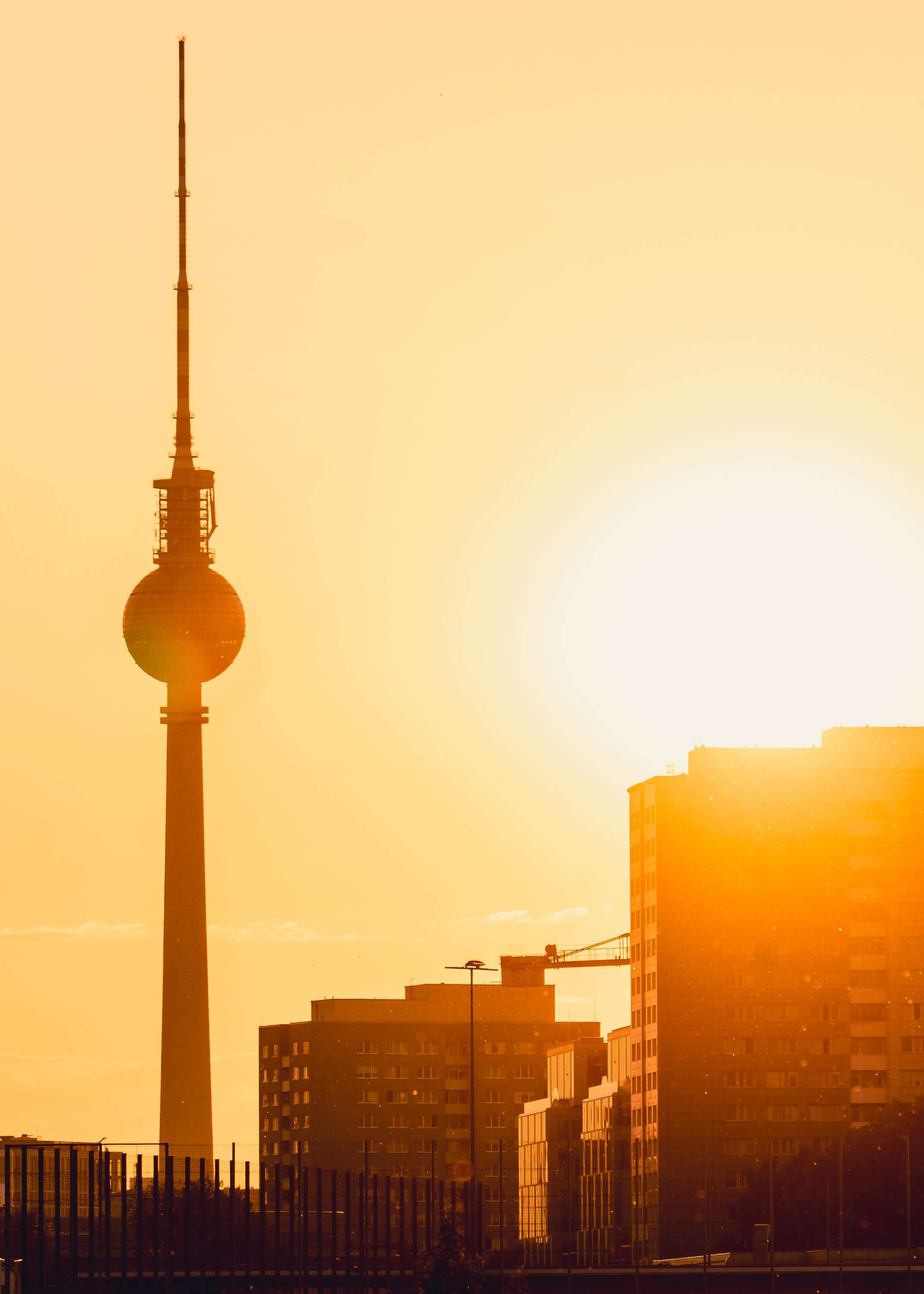 Berlin's television tower in dark silhouette against a hazy golden sky, the setting sun beside its sphere and office blocks glowing below.
