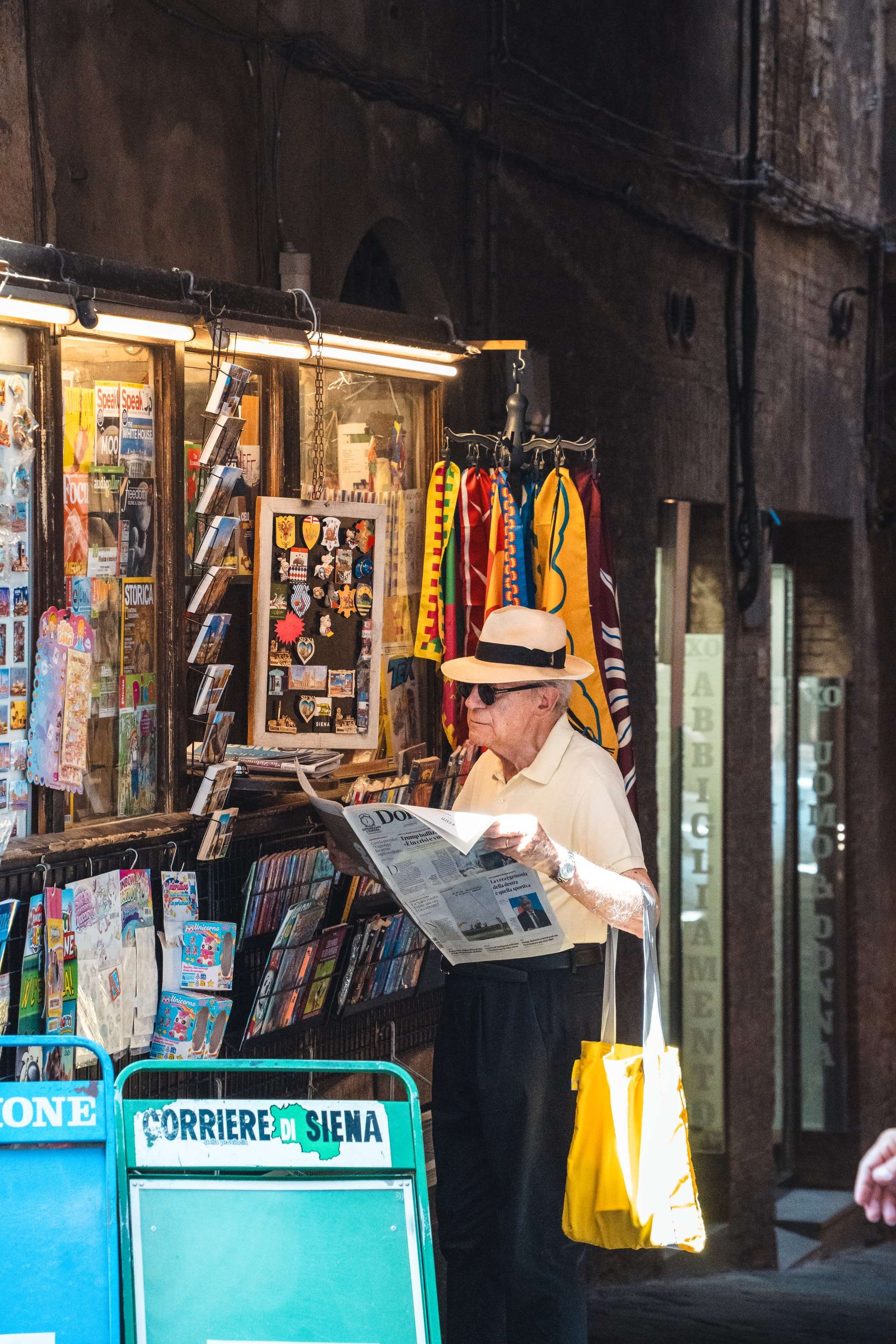 An elderly man in a Panama hat and sunglasses reads a newspaper at a newsstand in a narrow Siena street, a yellow bag on his arm and contrada flags hanging beside him.