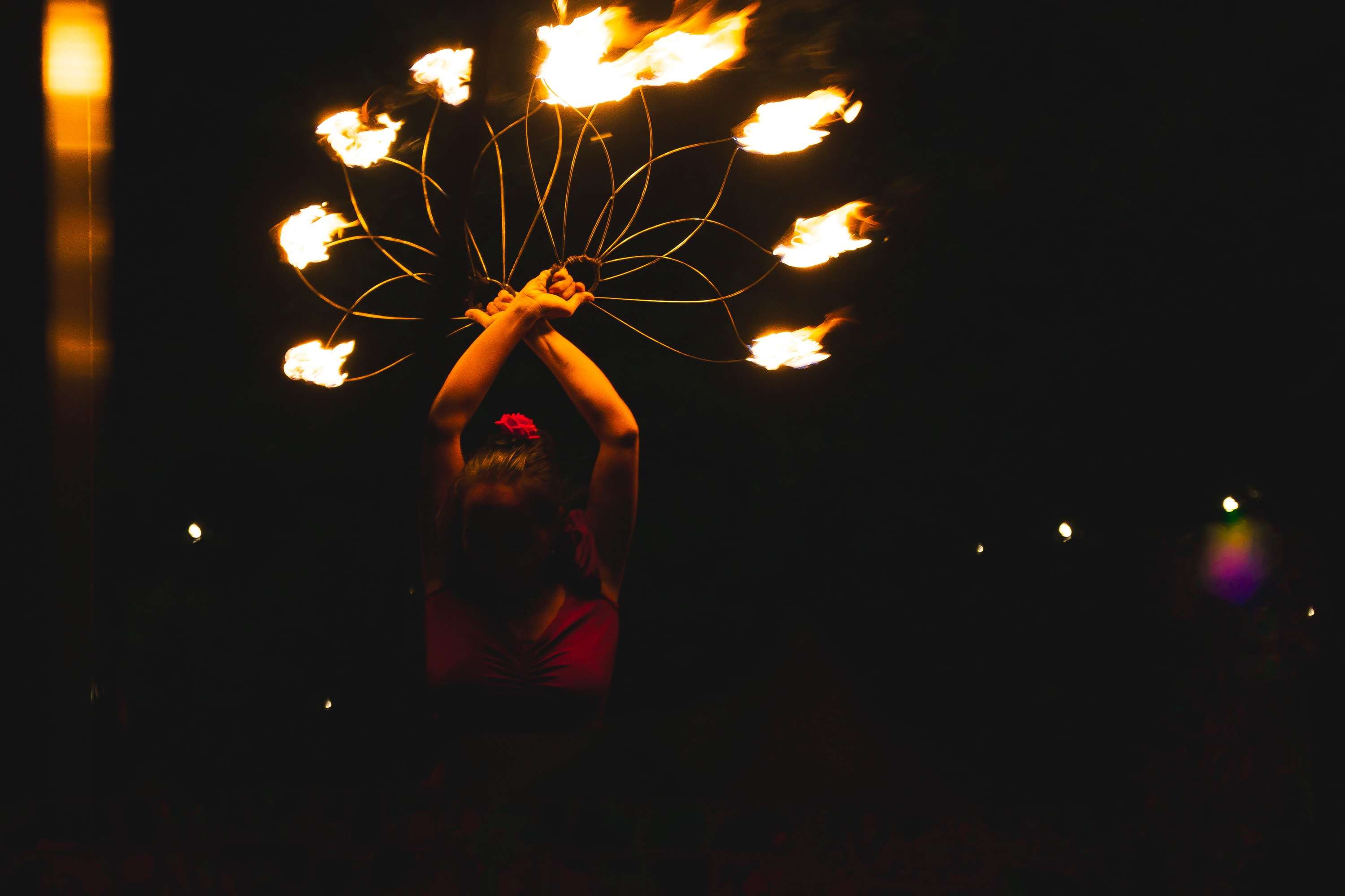 A fire dancer holds a fan of eight flaming wicks high over her head against a black night, her face lost in shadow.