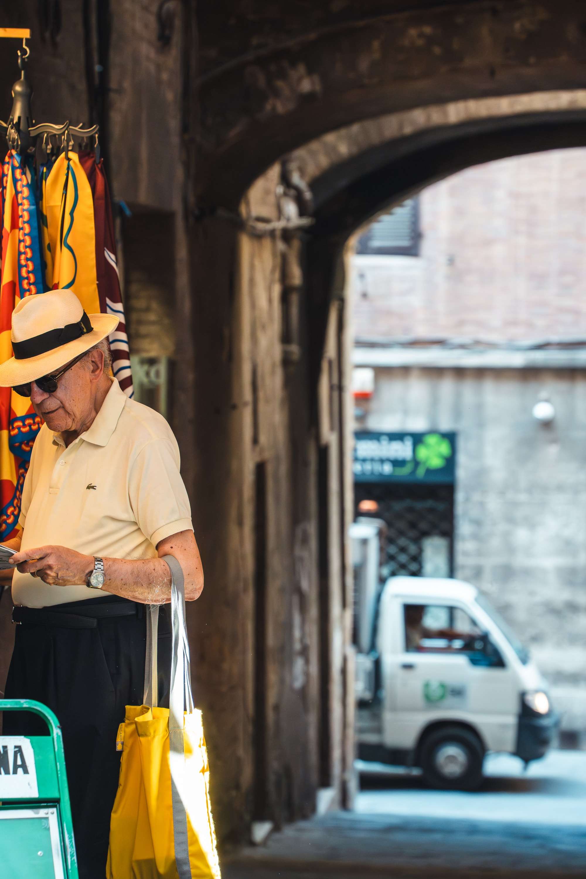 An elderly man in a Panama hat reads beside a stone archway in Siena, holding a yellow bag, a small delivery van parked in the sunny street beyond.