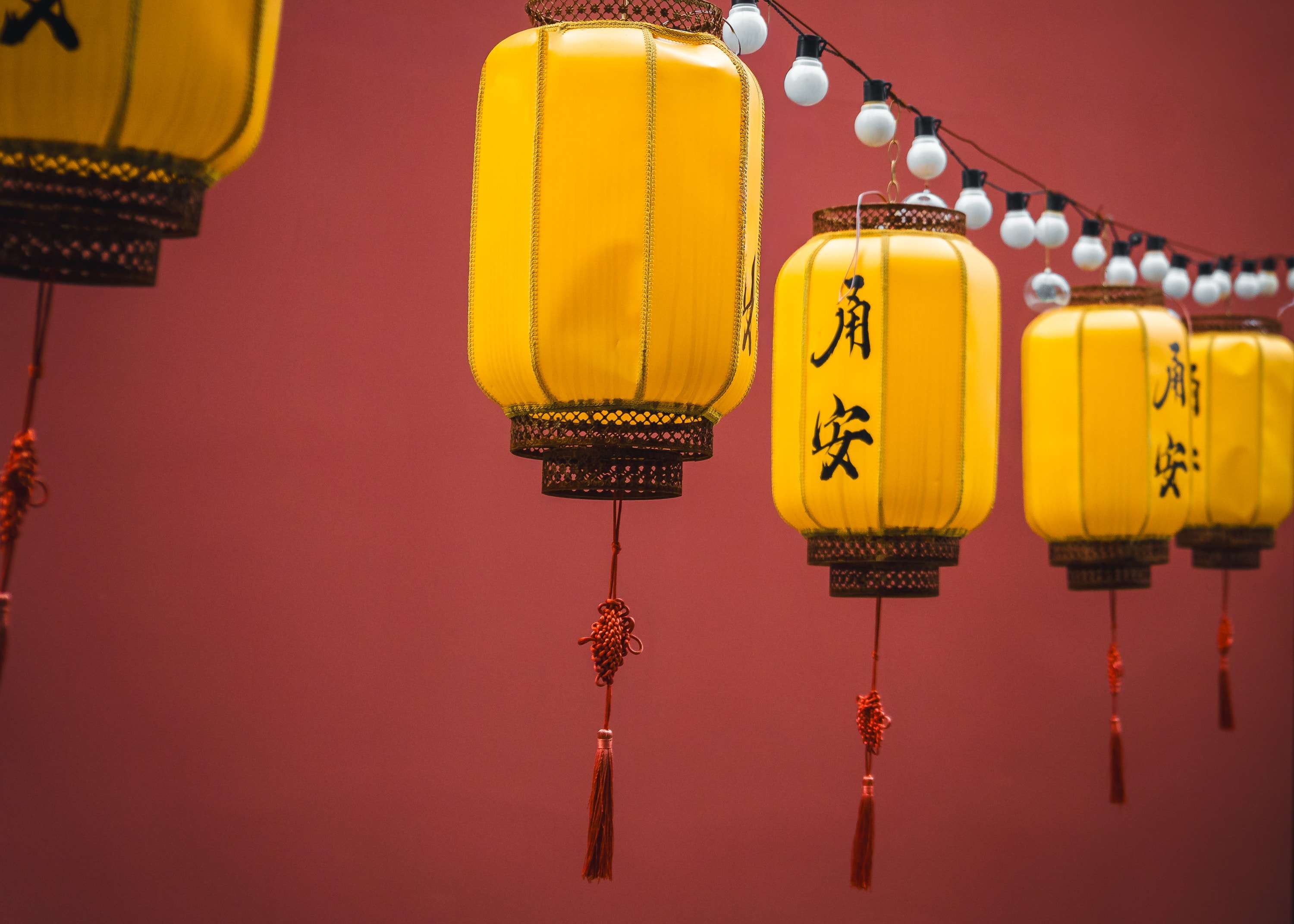 Yellow silk lanterns painted with the characters for peace and safety hang in a row against a red wall, with a string of white bulbs above.