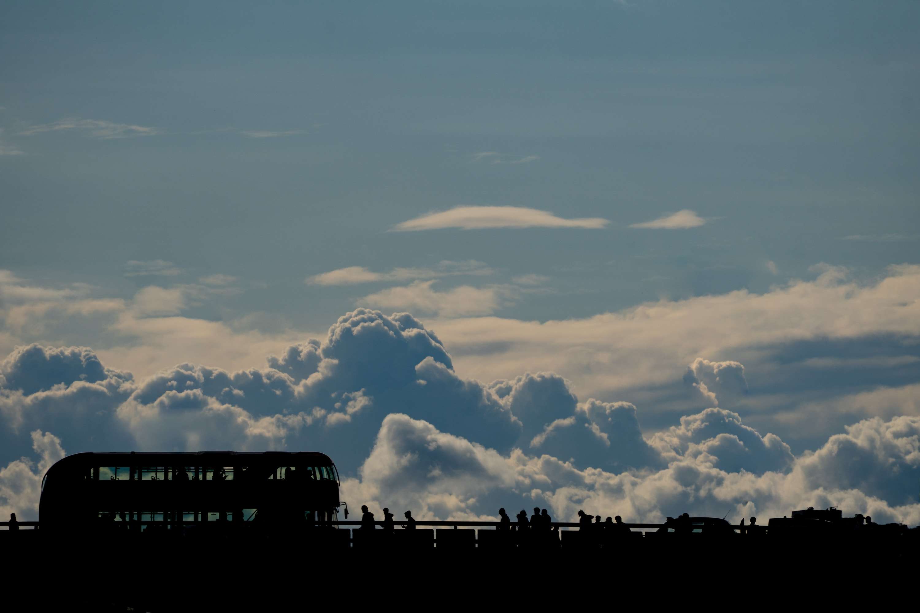 The black silhouette of a double-decker bus and a line of pedestrians along a bridge, below a bank of white cloud and a blue sky.