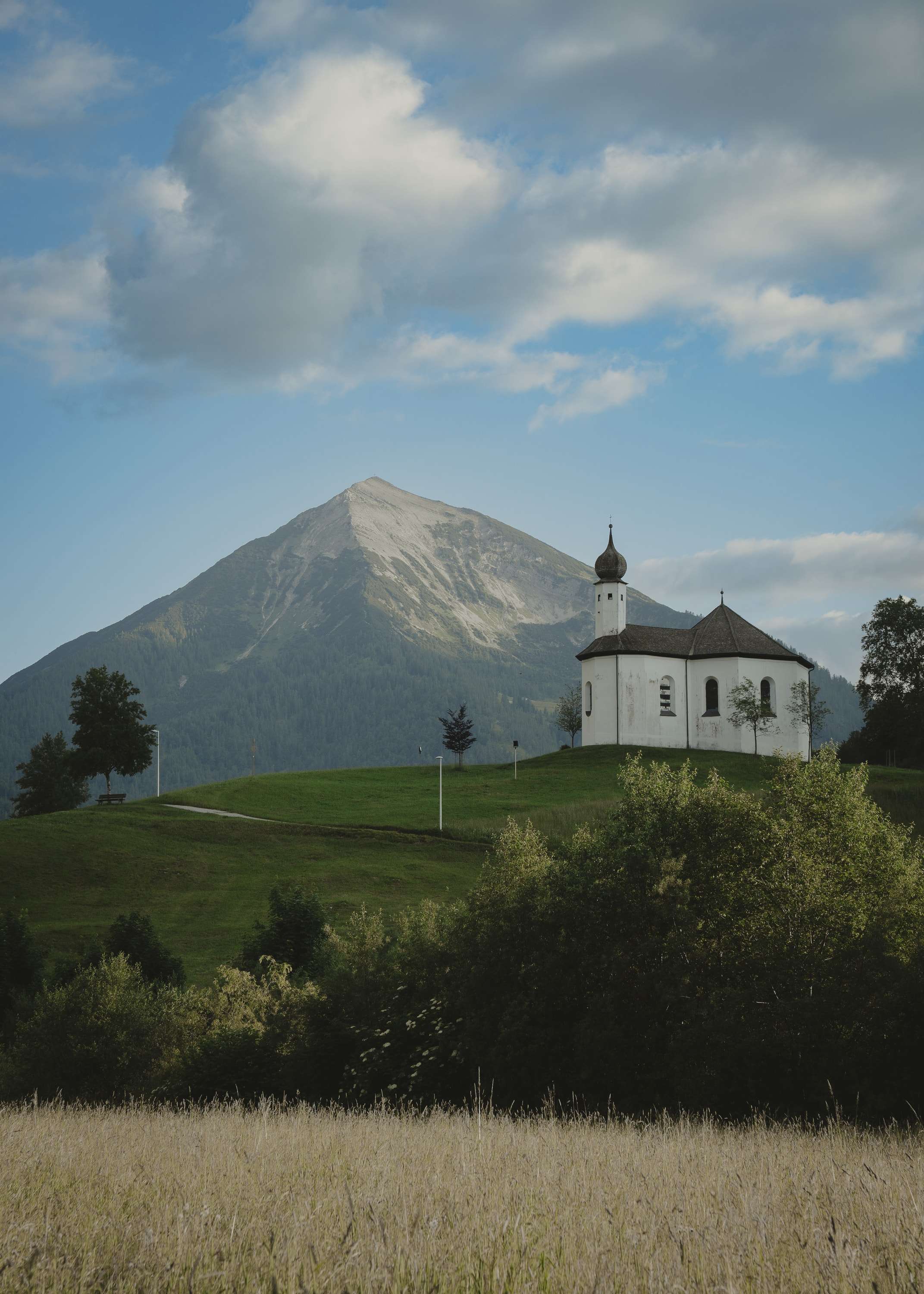 A small white chapel with an onion-domed tower sits on a green hill in front of a pointed grey mountain, with a field of pale grass in the foreground.