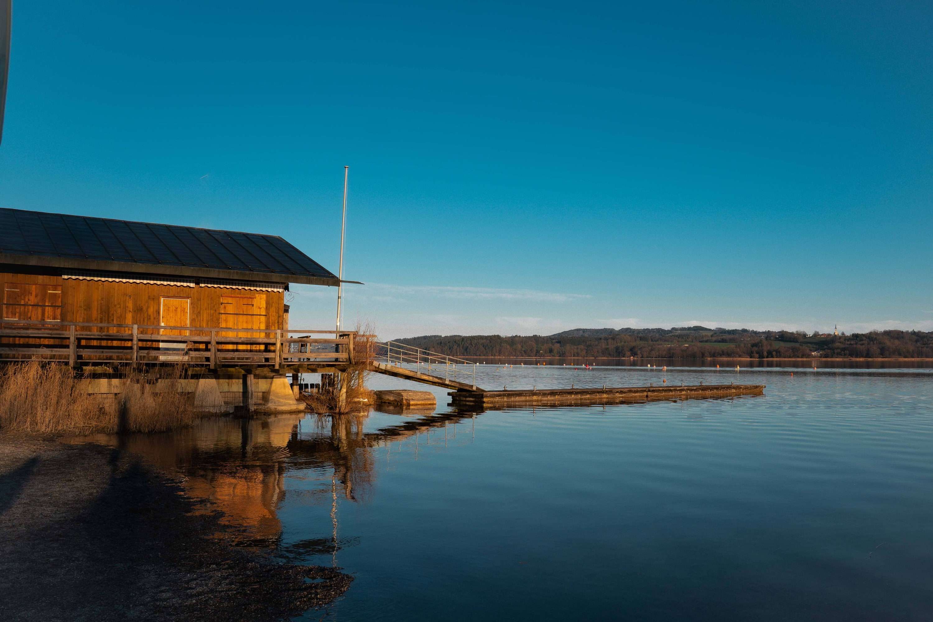 A wooden boathouse and jetty on a still lake in low sun, mirrored in the water, under a clear blue sky.
