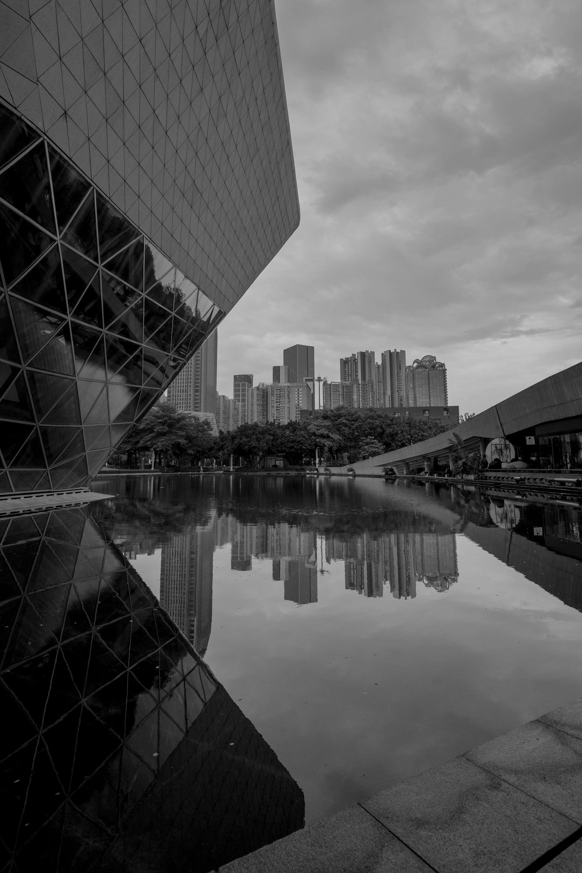 Black and white: the angled glass wall of the Guangzhou Opera House reflected in a still pool, with a line of tower blocks across the water.