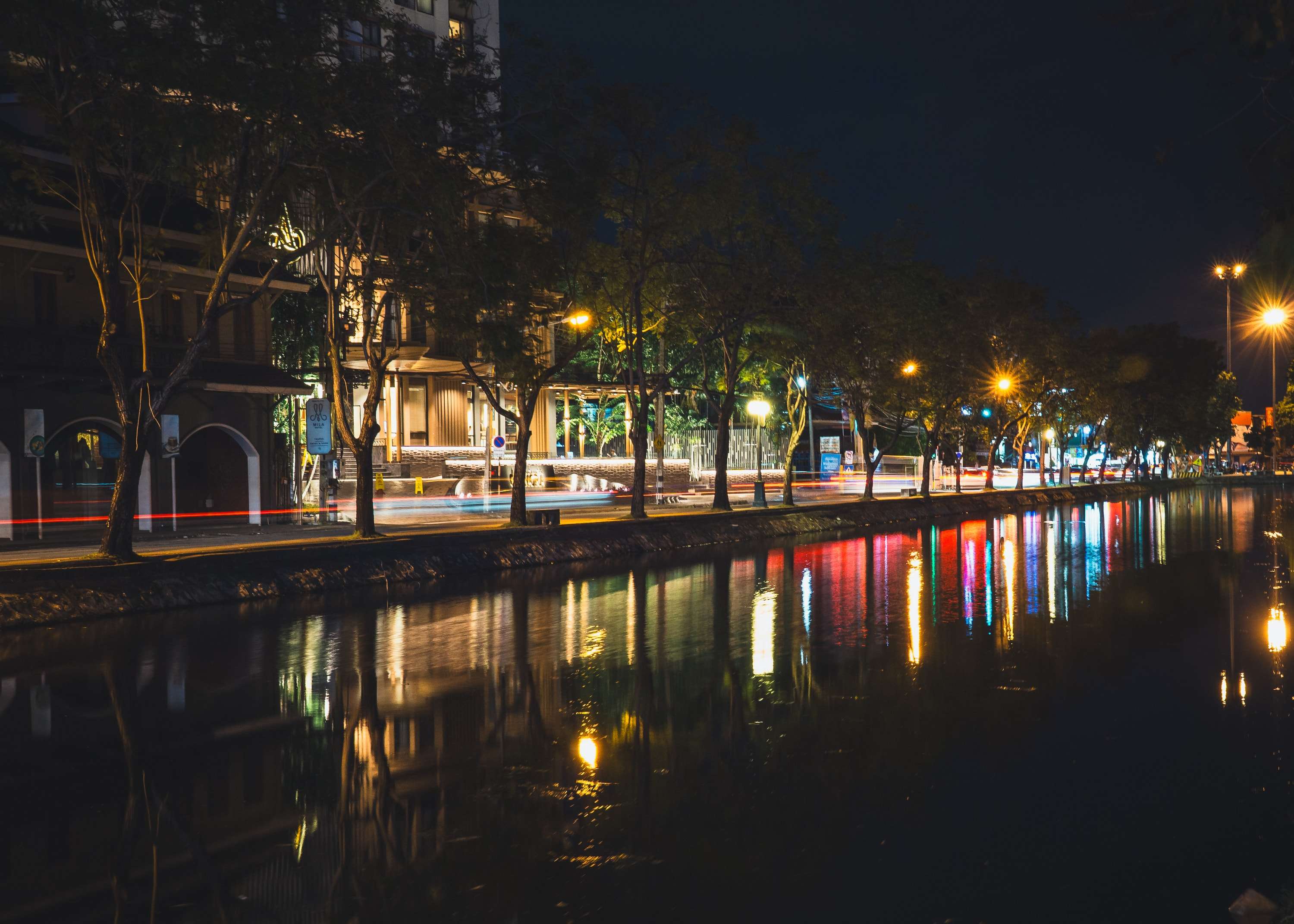 A still canal at night lined with trees and street lamps, the lights of shops and passing traffic reflected as coloured streaks in the water.