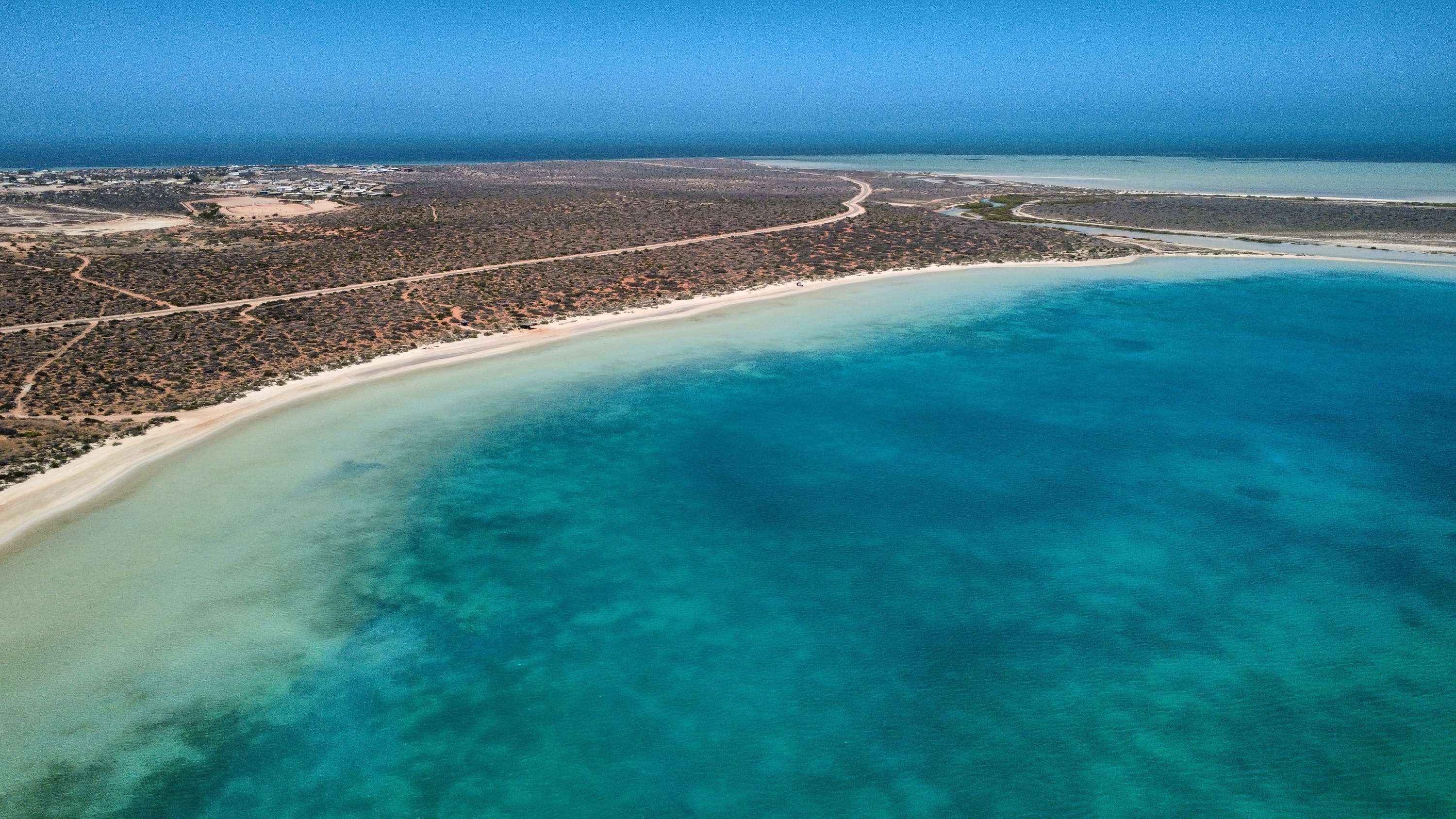 Aerial view of a curving white beach between turquoise shallow water and red scrubland, with a pale bay and a small town on the horizon.