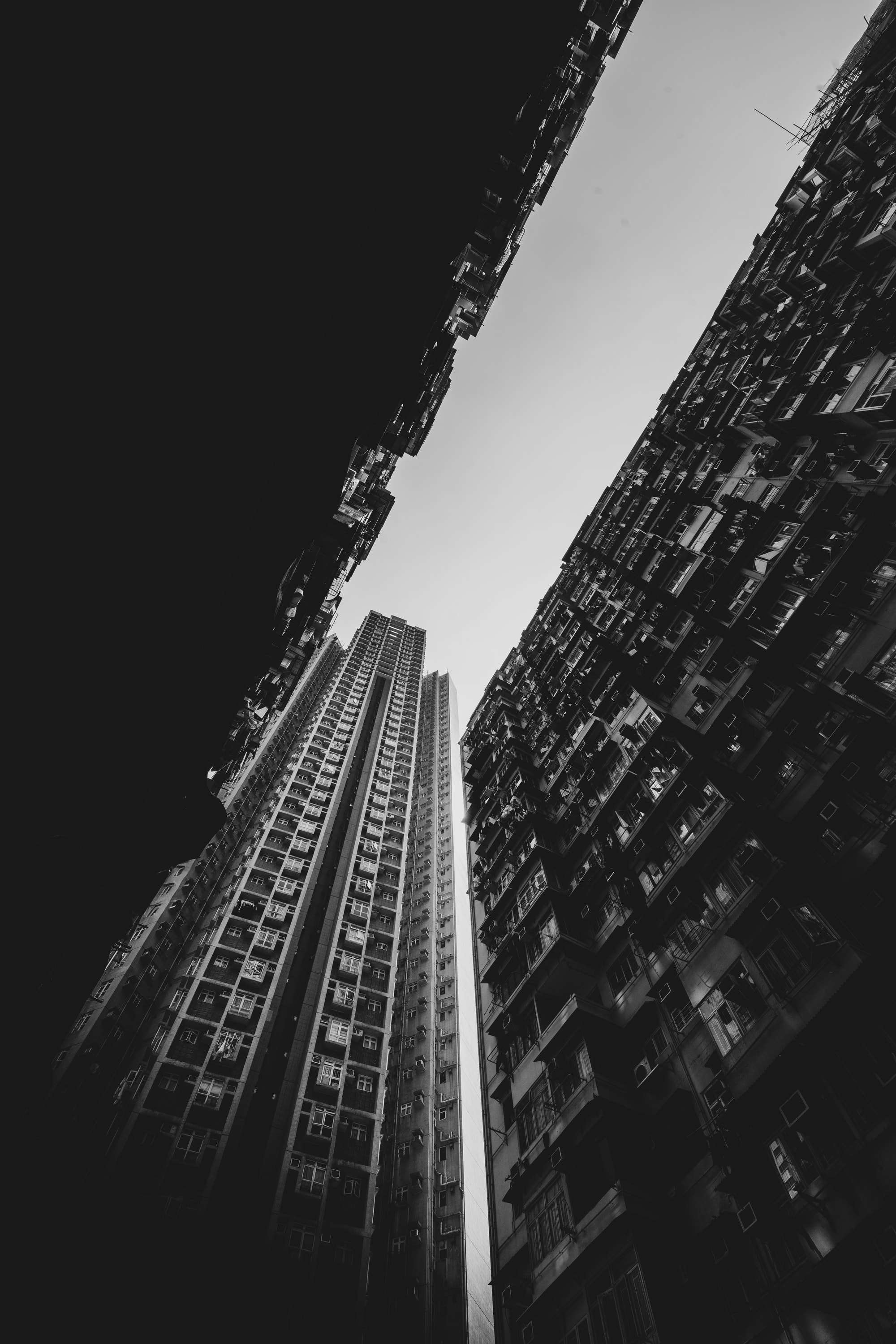 Black and white vertical view up between two walls of apartments, a narrow wedge of pale sky between them and a slender tower block in the gap.