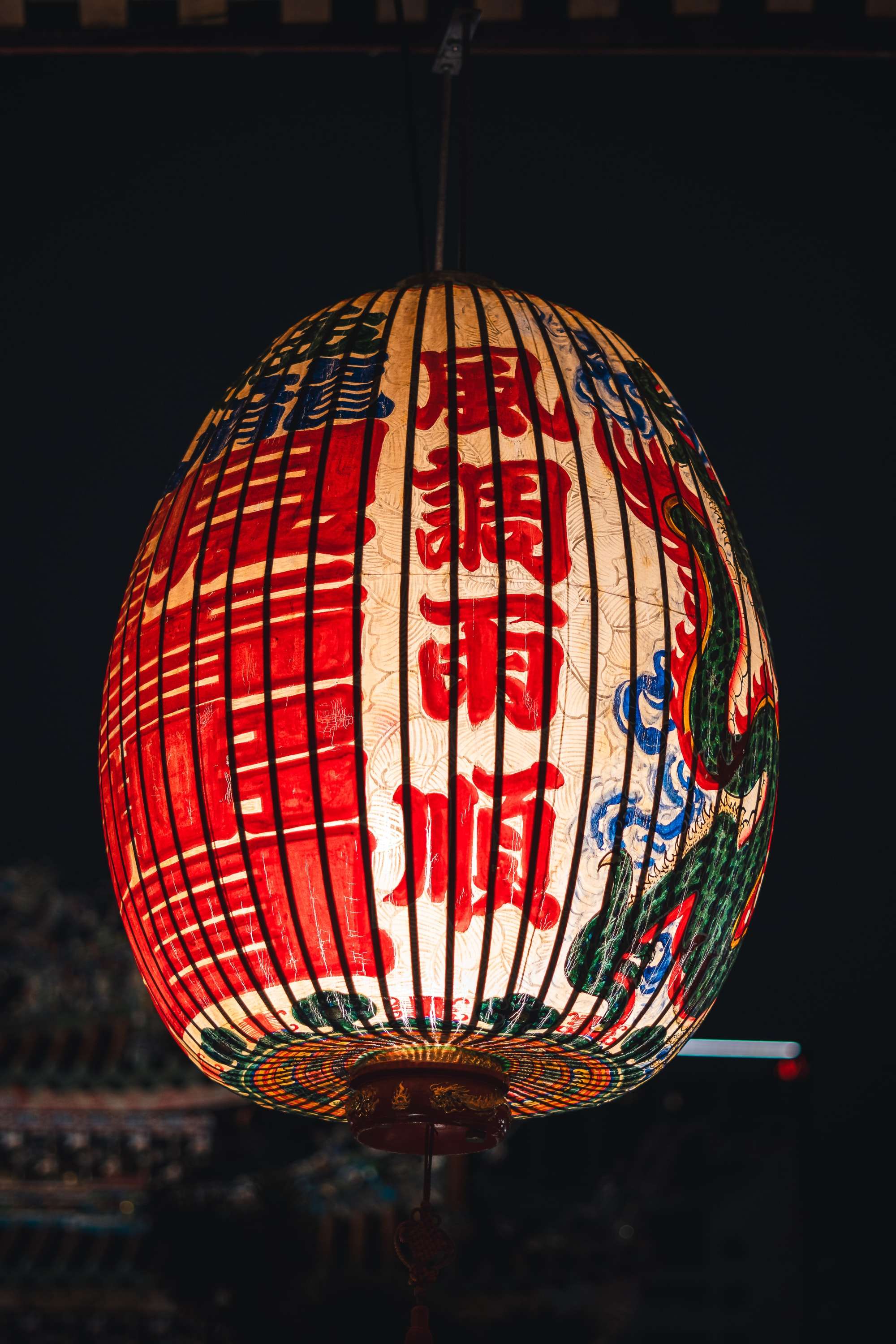 A large oval lantern with red characters and a green painted dragon hangs lit against the black night sky above a temple roof.