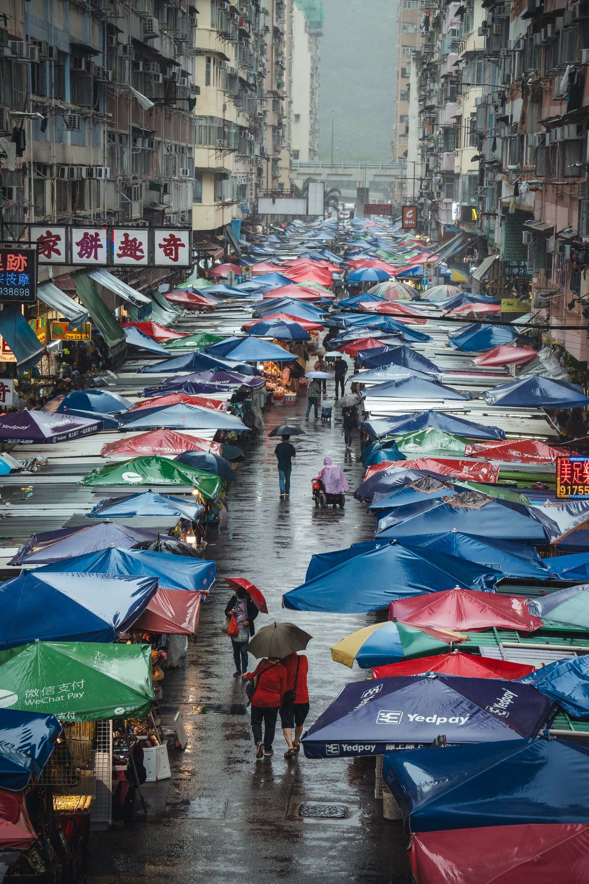 Looking down a street market in the rain: a long corridor of red, blue and green stall umbrellas between tall tenement blocks, people walking with umbrellas below.