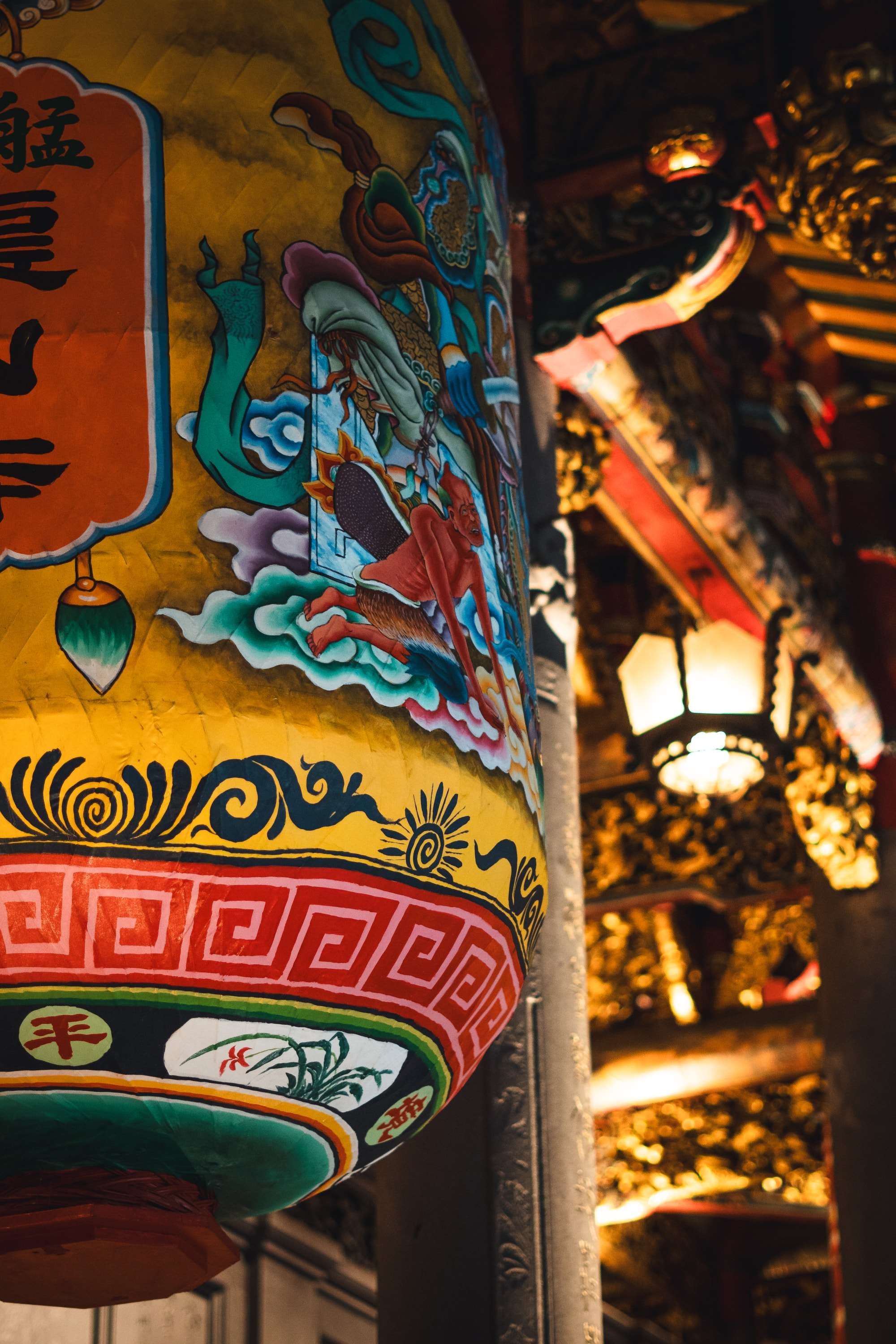 Close view of a large yellow temple lantern painted with a figure in blue and red robes and a red key pattern, lit beside a gilded wooden beam.