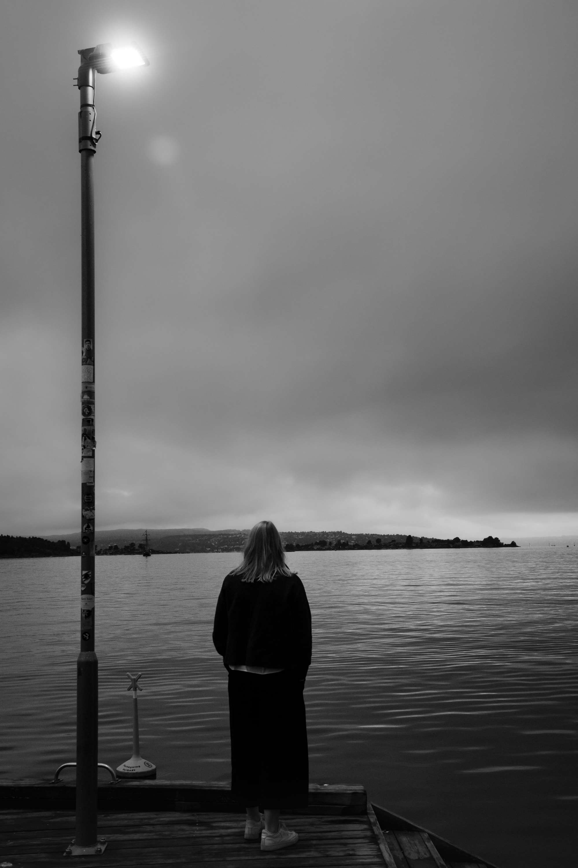 Black and white: a woman in a long dark coat stands at the end of a wooden pier beside a lit lamp post, looking out over the fjord under heavy cloud.