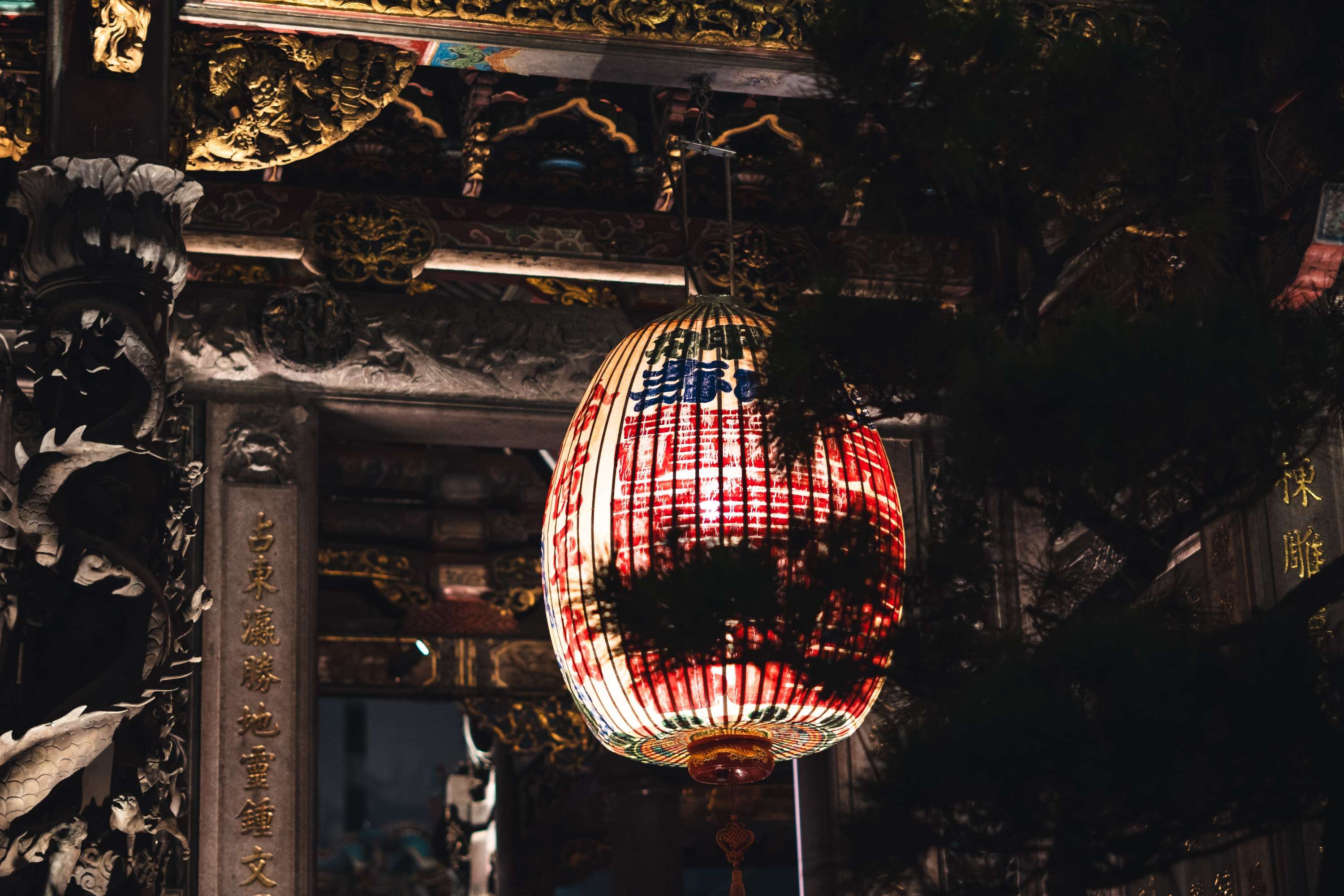 A red and white paper lantern glows under the carved and gilded eaves of Longshan Temple at night, with dark tree branches in front of it.